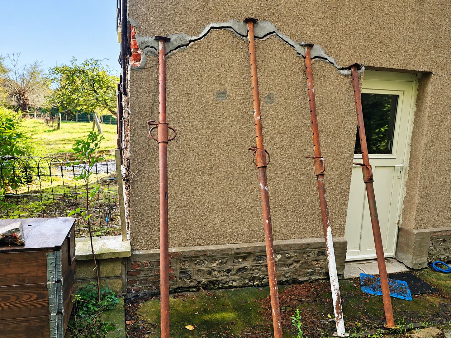 Un mur de maison en crépi présentant une large fissure en haut est soutenu par quatre étais métalliques rouillés, à côté d'une porte verte.