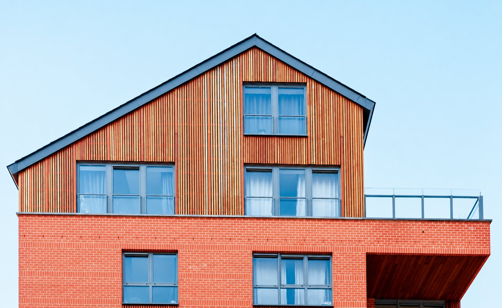 Façade d'un immeuble moderne associant brique rouge en partie basse et bardage en bois à lamelles verticales sous un toit à pignon, avec une terrasse à garde-corps vitré.